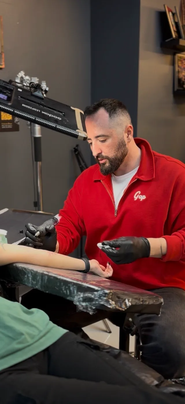 BORZ tattooing a client's forearm, wearing black gloves.