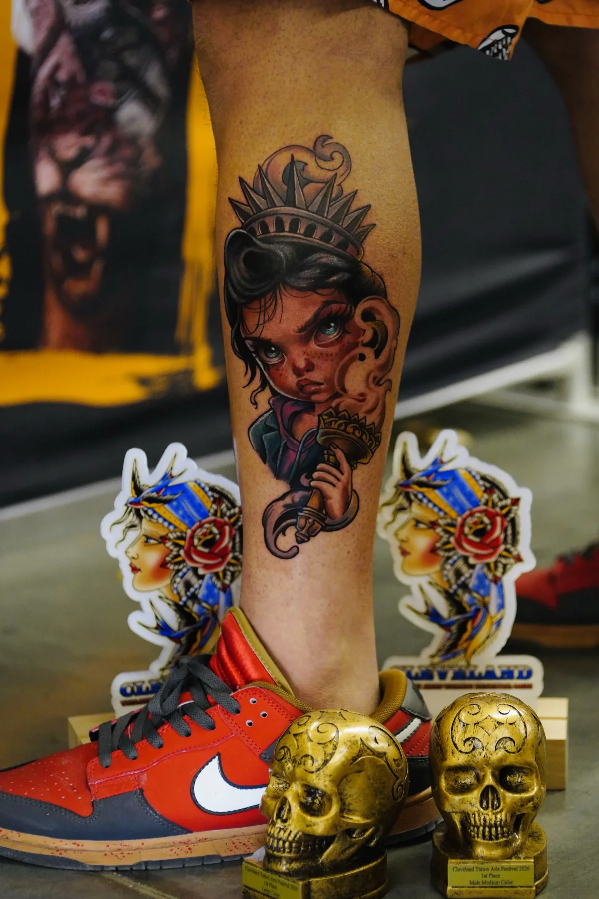 Neotraditional calf tattoo of a girl dressed as the Statue of Liberty, photographed next to two golden skull trophies.