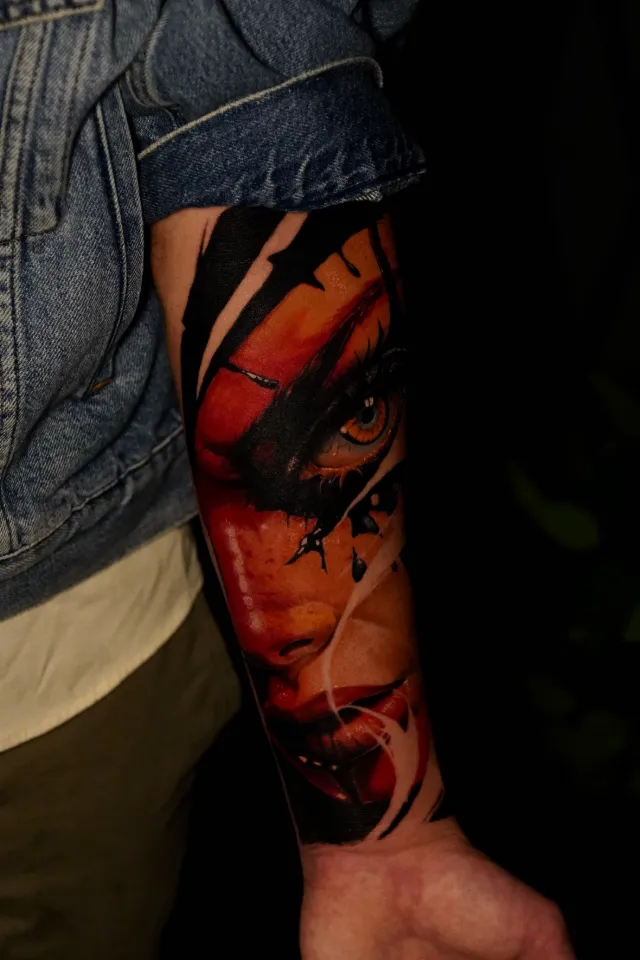 Color realism forearm tattoo of a woman's face in red and orange tones with an amber eye and black brushstrokes.