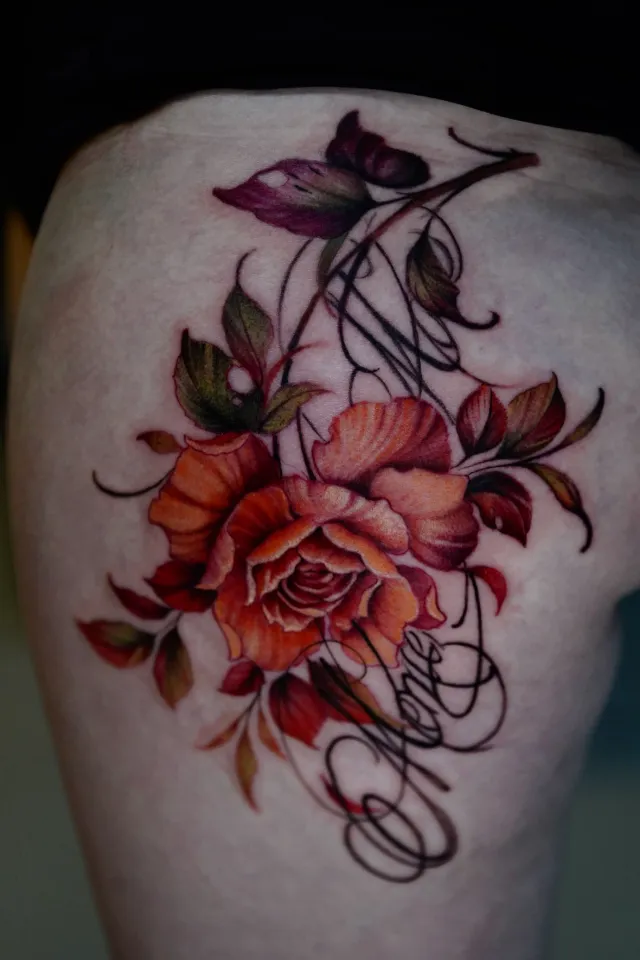 Tatuaje de realismo a color en la cadera: una rosa naranja con hojas rojas y verdes y letras caligráficas negras.