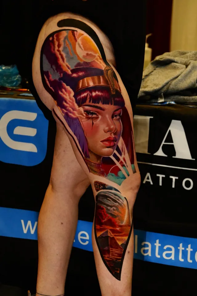 Tatuaje de realismo a color en la pierna: una reina egipcia con corona de cobra dorada y nubes violetas, con pirámides al atardecer en la pantorrilla.
