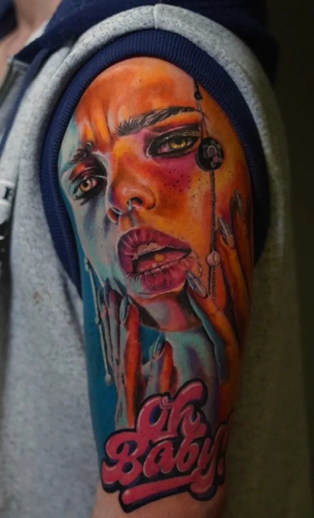 Tatuaje de realismo a color en el brazo: el rostro de una mujer en tonos naranjas y azules con las letras rosas "Oh Baby!".