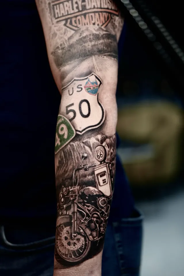 Tatuaje en negro y gris en el antebrazo: una motocicleta, un surtidor de gasolina de la Ruta 66 y las señales de la US 50 y la California 49.